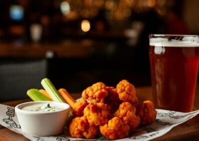 “Crispy Buffalo cauliflower wings coated in spicy orange sauce, served with ranch dip, celery sticks, and carrot slices on a white plate.”